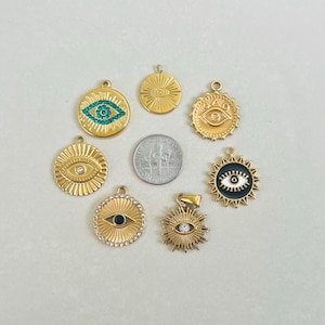 May include: Seven gold-toned circular eye-themed pendants arranged around a central US dime coin. The charms feature various designs including turquoise crystals, clear rhinestones, black enamel, and sunburst patterns, each with a small top loop for attachment.