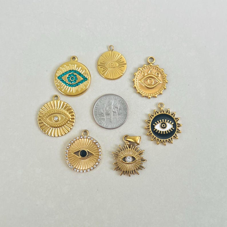 May include: Seven gold-toned circular eye-themed pendants arranged around a central US dime coin. The charms feature various designs including turquoise crystals, clear rhinestones, black enamel, and sunburst patterns, each with a small top loop for attachment.