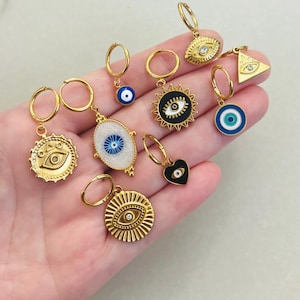 Op de afbeelding: Een set van negen gouden oorbellen met verschillende boze oogcharms. De charms omvatten een hart, een zon, een ovaal, een driehoek en een cirkel. De oorbellen liggen op een hand.