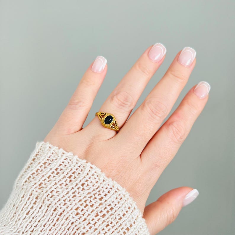 May include: A gold-colored ring with an oval, dark blue stone. The ring has an open filigree design around the stone. The ring is on a person's finger, with a light gray background.