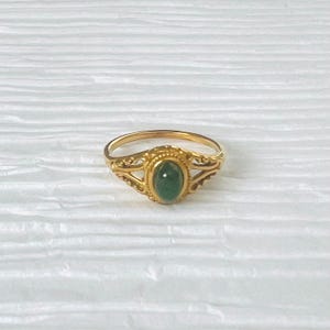 May include: A gold ring featuring an oval, emerald-green gemstone. The ring has an intricate, filigree design on the band and around the central stone, giving it a vintage or antique appearance.