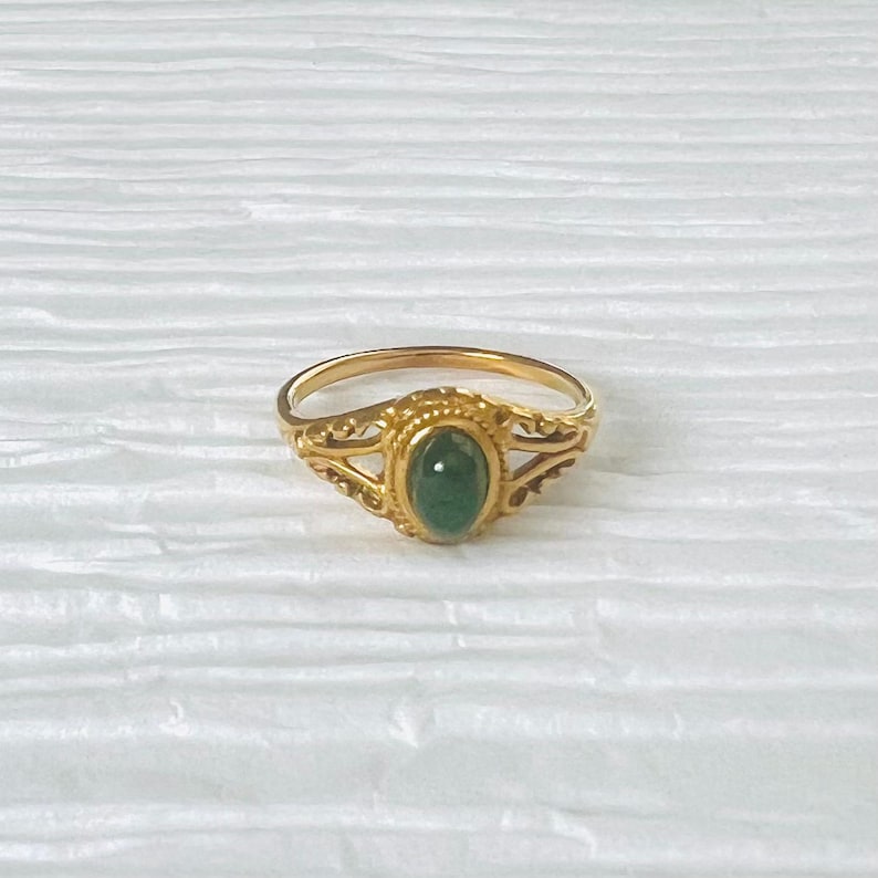 May include: A gold ring featuring an oval, emerald-green gemstone. The ring has an intricate, filigree design on the band and around the central stone, giving it a vintage or antique appearance.