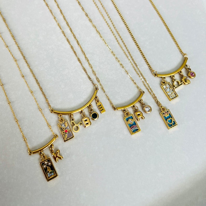 Peut inclure: Cinq colliers en or avec des breloques. Chaque collier est compos&eacute; d'une barre incurv&eacute;e avec des breloques qui pendent. Les breloques comprennent des cartes de tarot, des lettres et des chiffres. Les breloques sont color&eacute;es et ont un style vintage.