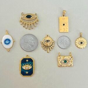 May include: A collection of gold-toned metal pendants featuring various eye motifs, including sunburst, rectangular, and oval shapes with enamel and crystal accents. Two US quarters are included for scale, each measuring 0.955 inches in diameter.