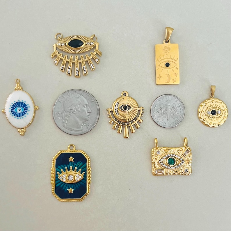 May include: A collection of gold-toned metal pendants featuring various eye motifs, including sunburst, rectangular, and oval shapes with enamel and crystal accents. Two US quarters are included for scale, each measuring 0.955 inches in diameter.