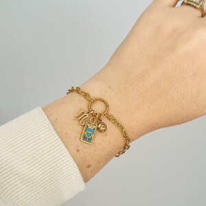 May include: Gold chain bracelet with a charm featuring a blue and gold tarot card with a heart and sun design, a gold charm with a crystal, and a gold charm with the letter "M".