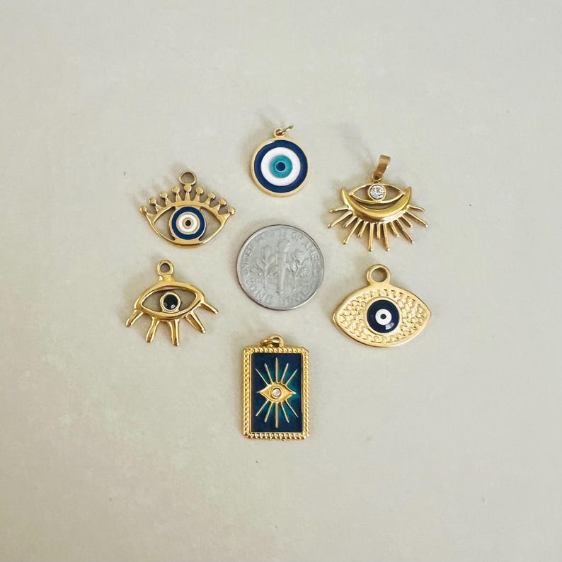 May include: Six gold-tone metal charms featuring evil eye motifs in blue, white, and black enamel, arranged around a silver-colored US dime for scale. The collection includes circular, rectangular, and stylized eye-shaped pendants with metal rays.