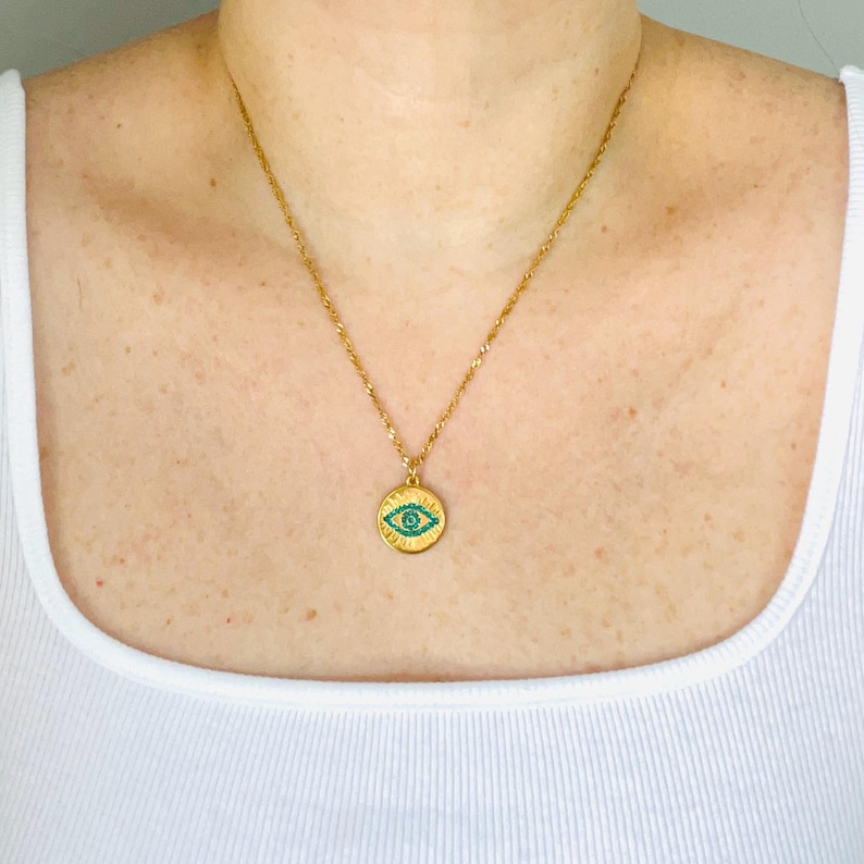 May include: A gold-toned chain necklace featuring a circular pendant with a central eye motif. The eye design is detailed with small, vibrant turquoise-colored stones set against a textured gold background, creating a distinct focal point.