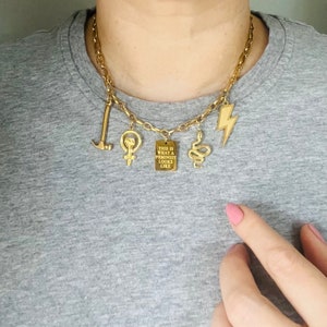 May include: Gold chain necklace with multiple charms, including a hammer, a fist, a snake, a lightning bolt, and a rectangular charm with the text "This is what a feminist looks like".