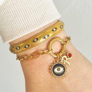 May include: Gold bracelet with a charm featuring a black and white eye design. The bracelet is adorned with a chain and a small red charm with the letter "R".