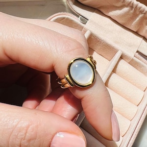 May include: A gold ring with a large, oval-shaped, white gemstone. The ring is set in a gold band with a textured design.