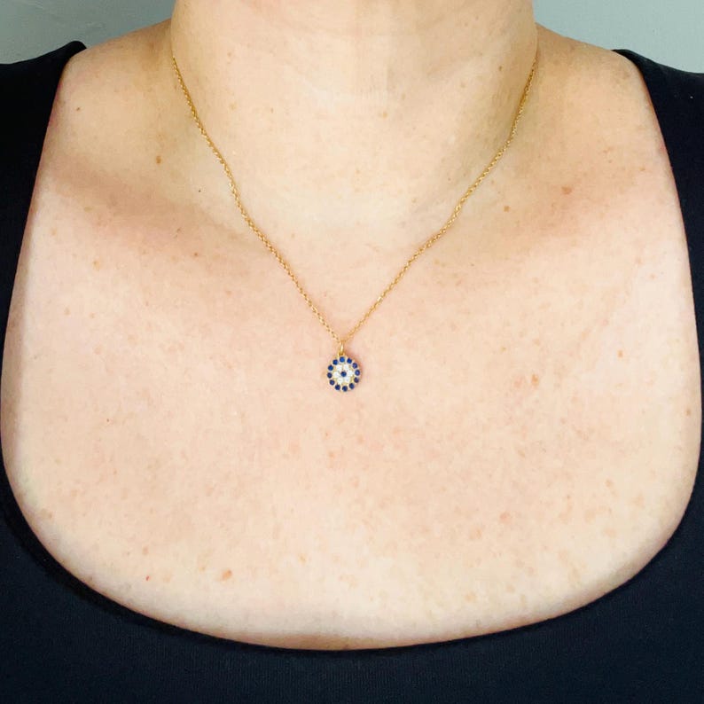 May include: A gold-toned chain necklace featuring a small, circular pendant. The pendant displays a central dark blue stone surrounded by a ring of tiny white stones, creating a circular eye-like pattern that hangs centered on the delicate chain.