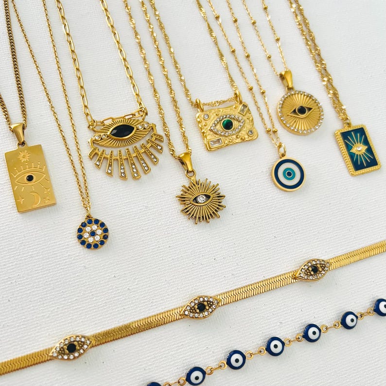 May include: A collection of gold-toned necklaces and bracelets featuring various evil eye motifs. The jewelry includes rectangular, circular, and sunburst pendants with blue, black, and clear crystal accents, alongside two flat herringbone chains.