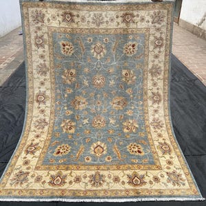 Könnte beinhalten: Ein rechteckiger, handgeknüpfter Teppich mit einem blau-grauen Mittelfeld, verziert mit floralen Mustern in Beige, Braun und Gold. Der Rand weist ein ergänzendes Design auf. Der Teppich wird auf einer schwarzen Oberfläche präsentiert.
