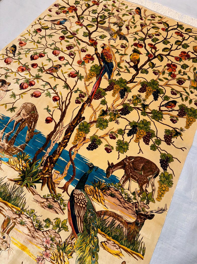 Op de afbeelding: Een cr&egrave;mekleurig tapijt met een gedetailleerd ontwerp van een boom met vogels, druiven en herten. De boom wordt omringd door een blauwe rivier en een pauw. Het tapijt heeft een vintage esthetiek.