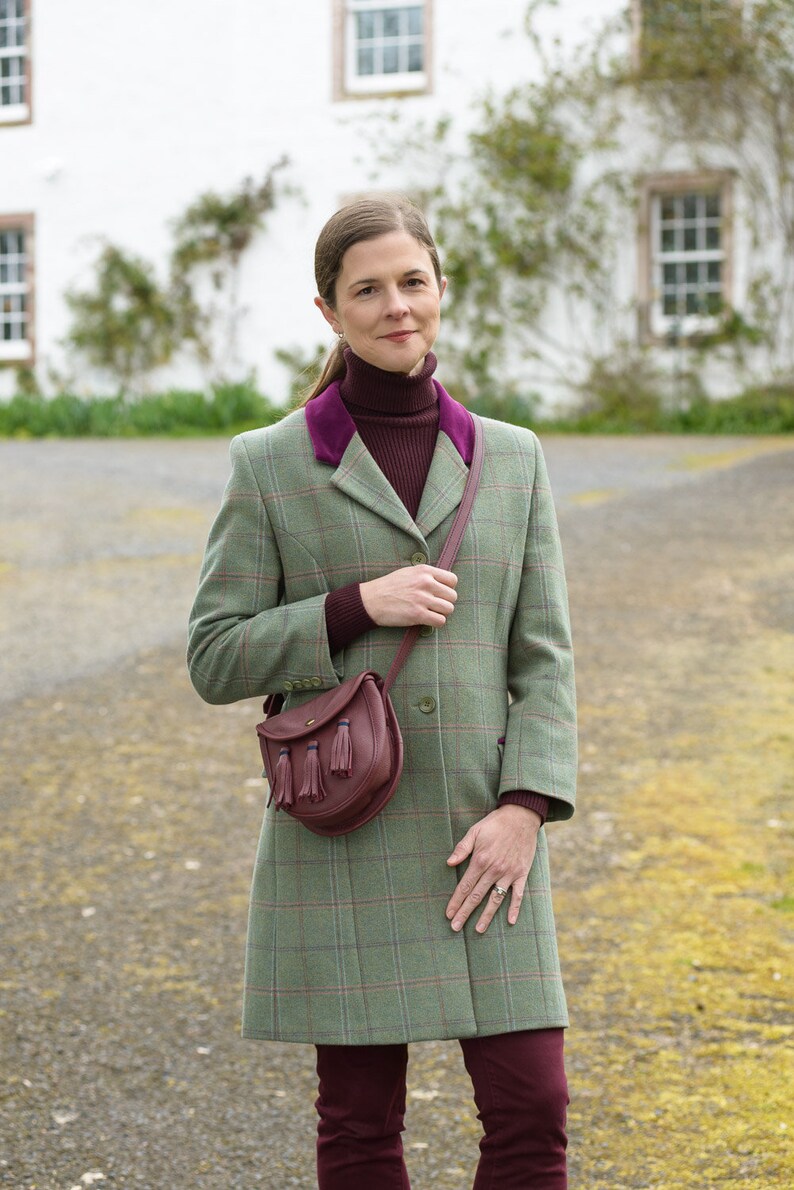 Pu&ograve; includere: Un cappotto in tweed a quadri verde e marrone con un colletto in velluto bordeaux e una borsa in pelle marrone con nappe. Il cappotto ha una chiusura con bottoni ed &egrave; indossato sopra un maglione a collo alto bordeaux.