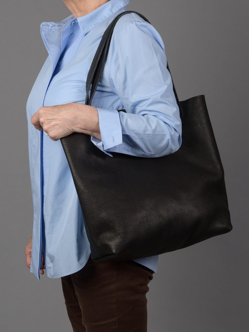 May include: A person carrying a black leather tote bag with a light blue button-down shirt and brown pants.