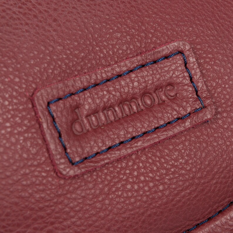 Pu&ograve; includere: Primo piano di una borsa in pelle bordeaux con un'etichetta cucita blu che recita "dunmore".