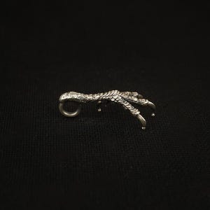 May include: A silver-toned pendant shaped like a bird's claw. The claw features intricate scale detailing and three extended talons. A small loop is present at the top for attaching a chain. The background is a plain black.