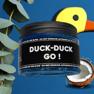 May include: A black jar with a blue slime inside. The jar has a black label with white text that reads "DUCK-DUCK GO!"  The jar is sitting on a blue background with a yellow duck and a coconut.