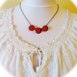 May include: A delicate necklace with three red flower charms on a black chain. The necklace is displayed on a white blouse with a ruffled neckline.