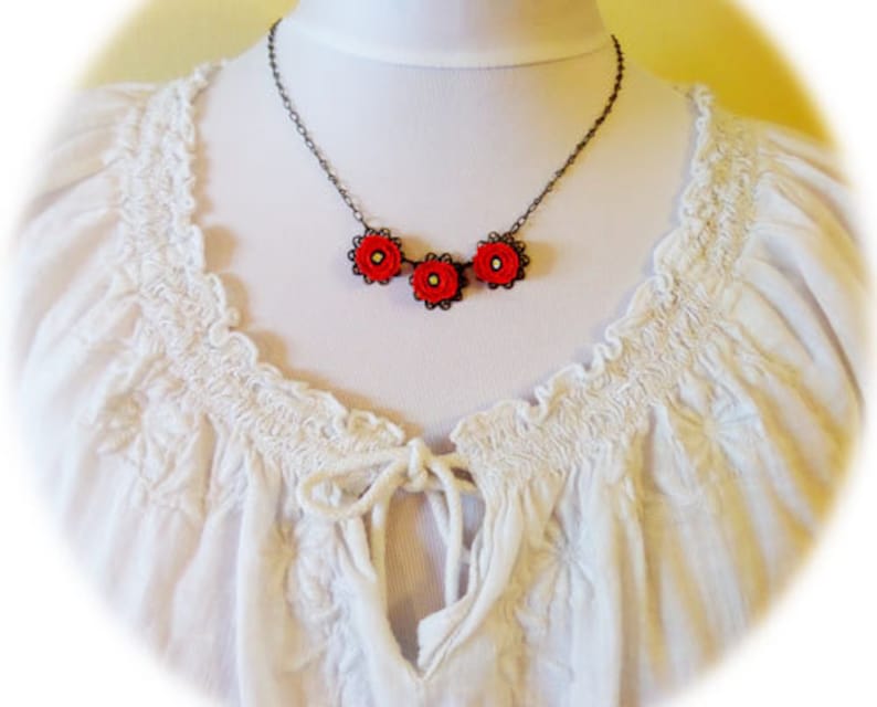 May include: A delicate necklace with three red flower charms on a black chain. The necklace is displayed on a white blouse with a ruffled neckline.