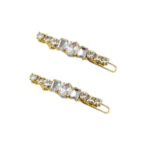 May include: Two gold-toned hair barrettes, each adorned with a line of clear, sparkling rhinestones. The rhinestones vary in size and shape, creating a glamorous accessory. The barrettes are set against a white background.