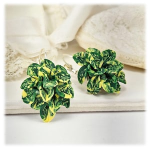 May include: A pair of handmade earrings featuring a cluster of green and yellow leaves. The earrings have a silver-colored hook closure. The leaves are detailed with a marbled effect, creating a naturalistic look. The earrings are set against a white background.