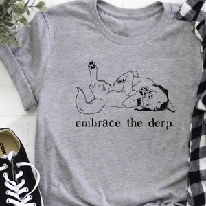 Puede incluir: Camiseta gris con una ilustración en blanco y negro de un perro acostado de espaldas con las patas en el aire. El texto "embrace the derp." está impreso debajo del perro.