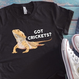 Può includere: Maglietta nera con una grafica bianca di un drago barbuto e il testo "GOT CRICKETS?"