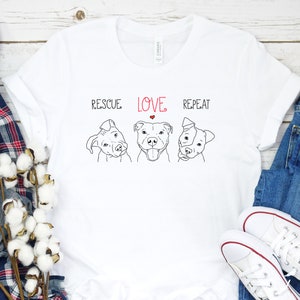 Puede incluir: Camiseta blanca con un dibujo en l&iacute;nea negra de tres perros pitbull. El texto "Rescue Love Repeat" est&aacute; impreso sobre los perros. Un coraz&oacute;n rojo est&aacute; en la palabra "Love".