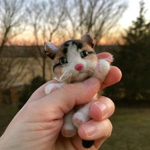 Op de afbeelding: Een klein, handgevoeld kattenbeeldje met bruin, wit en zwart bont. De kat heeft groene ogen en een roze neus. Het beeldje wordt in de hand van een persoon gehouden.