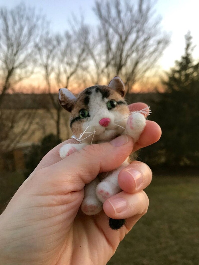 Op de afbeelding: Een klein, handgevoeld kattenbeeldje met bruin, wit en zwart bont. De kat heeft groene ogen en een roze neus. Het beeldje wordt in de hand van een persoon gehouden.