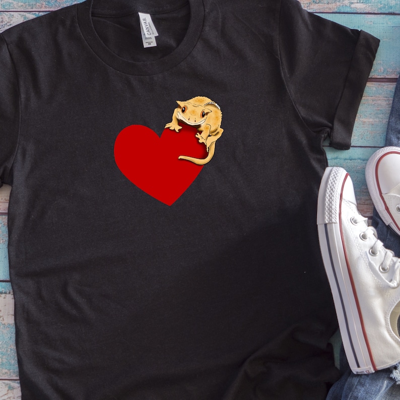 May include: Black t-shirt with a red heart and a small orange gecko on it. The gecko is holding onto the heart with its front legs.