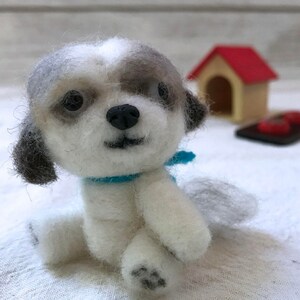 Op de afbeelding: Een klein, wit en grijs vilten hondenspeelgoed met een blauwe sjaal. De hond zit met zijn hoofd gekanteld en kijkt naar de camera.