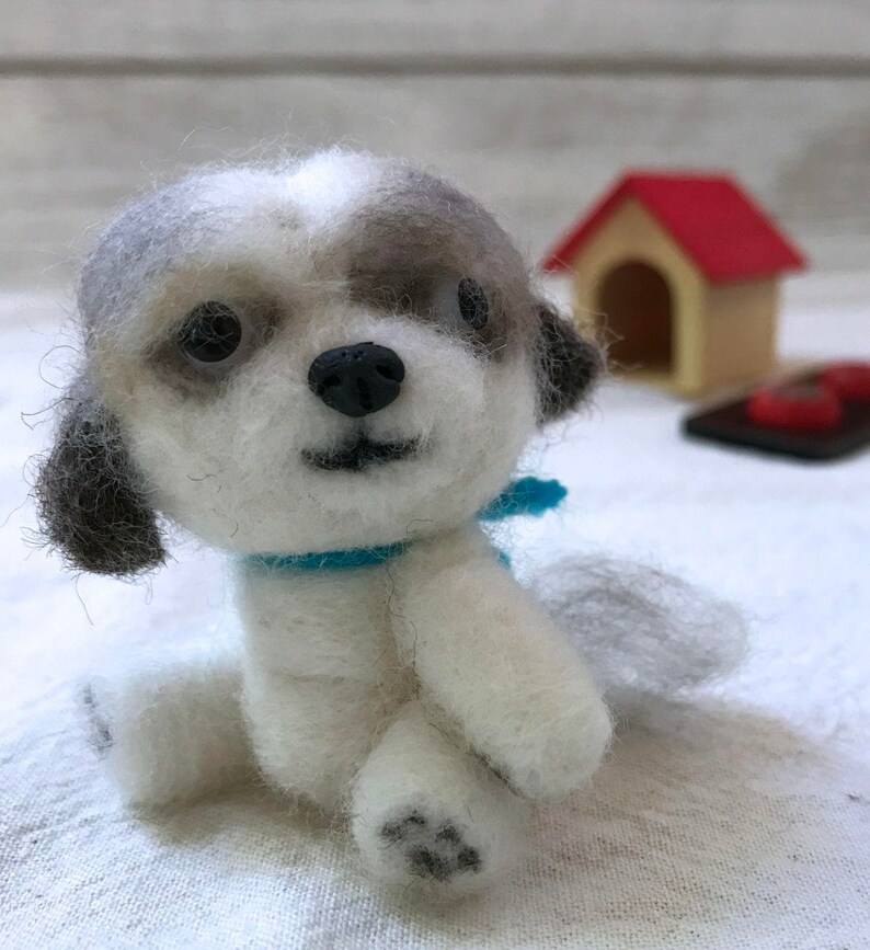 Op de afbeelding: Een klein, wit en grijs vilten hondenspeelgoed met een blauwe sjaal. De hond zit met zijn hoofd gekanteld en kijkt naar de camera.
