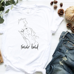 Puede incluir: Camiseta blanca con un dibujo en l&iacute;nea negra de una persona y un perro con el texto "Forever loved" debajo.
