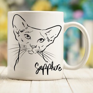 May include: White ceramic mug with a black line drawing of a Siamese cat with the name "Sapphire" written underneath.
