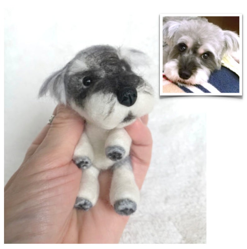 Op de afbeelding: Een klein, met de hand geviltd miniatuur schnauzer speelgoed. Het speelgoed is grijs en wit met zwarte ogen en een zwarte neus. Het zit met zijn poten in de lucht.