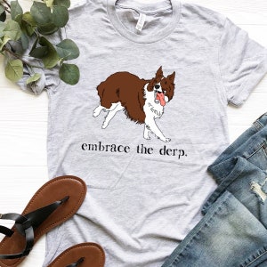 May include: A grey t-shirt featuring an illustration of a brown and white border collie dog. The dog is sticking its tongue out and has a playful expression. The text "embrace the derp." is printed below the dog.
