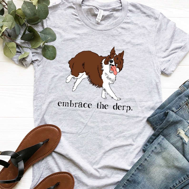 May include: A grey t-shirt featuring an illustration of a brown and white border collie dog. The dog is sticking its tongue out and has a playful expression. The text "embrace the derp." is printed below the dog.