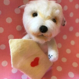 Op de afbeelding: Een witte knuffelhond die een klein envelopje vasthoudt met een rood hartje erop. De hond zit op een roze en witte stippenachtergrond.