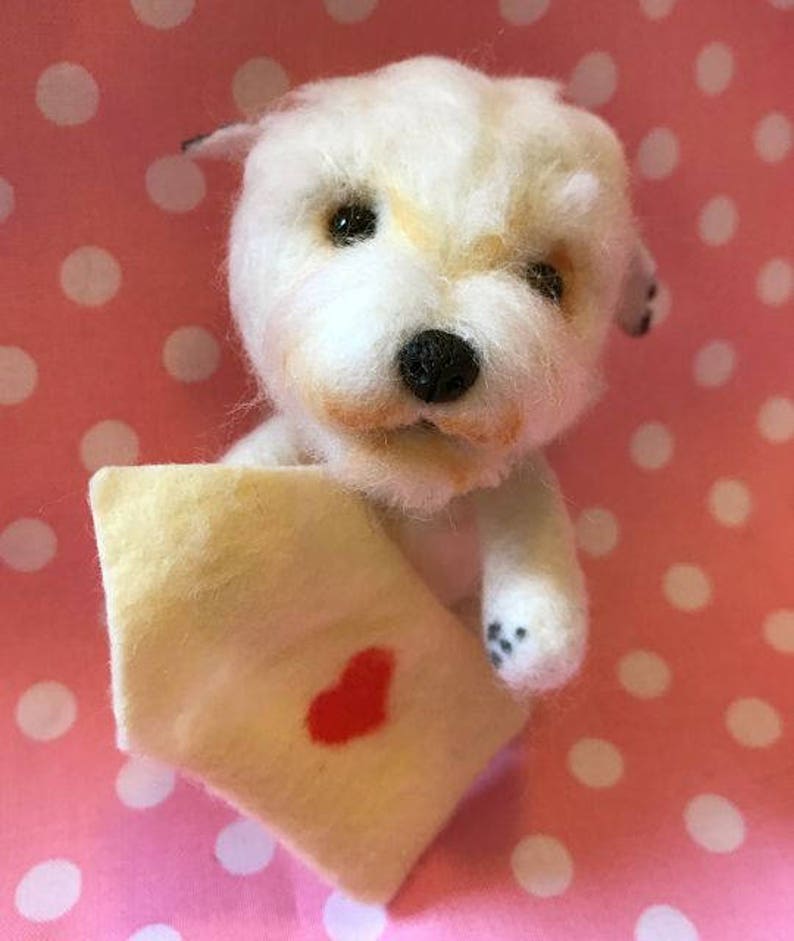 Op de afbeelding: Een witte knuffelhond die een klein envelopje vasthoudt met een rood hartje erop. De hond zit op een roze en witte stippenachtergrond.