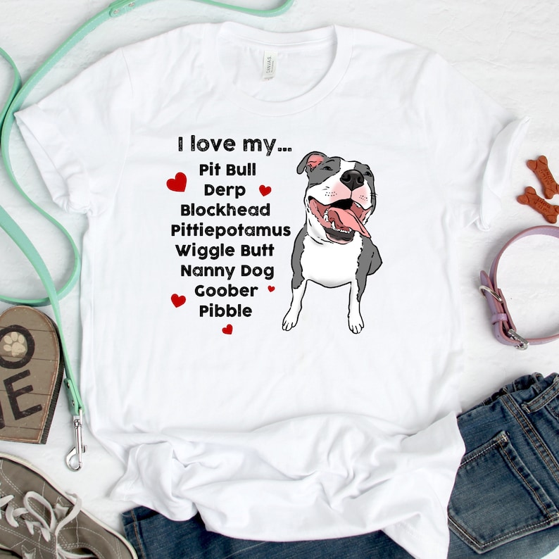 Op de afbeelding: Wit T-shirt met een cartoon afbeelding van een pit bull en de tekst "Ik hou van mijn... Pit Bull Derp Blockhead Pittiepotamus Wiggle Butt Nanny Dog Goober Pibble".