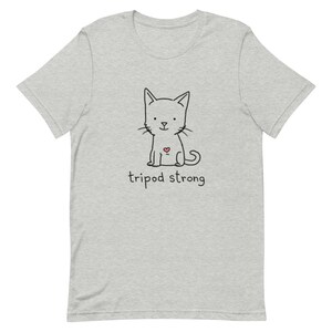 May include: A gray t-shirt with a black and white illustration of a cat with a red heart on its chest. The text "tripod strong" is printed below the cat.