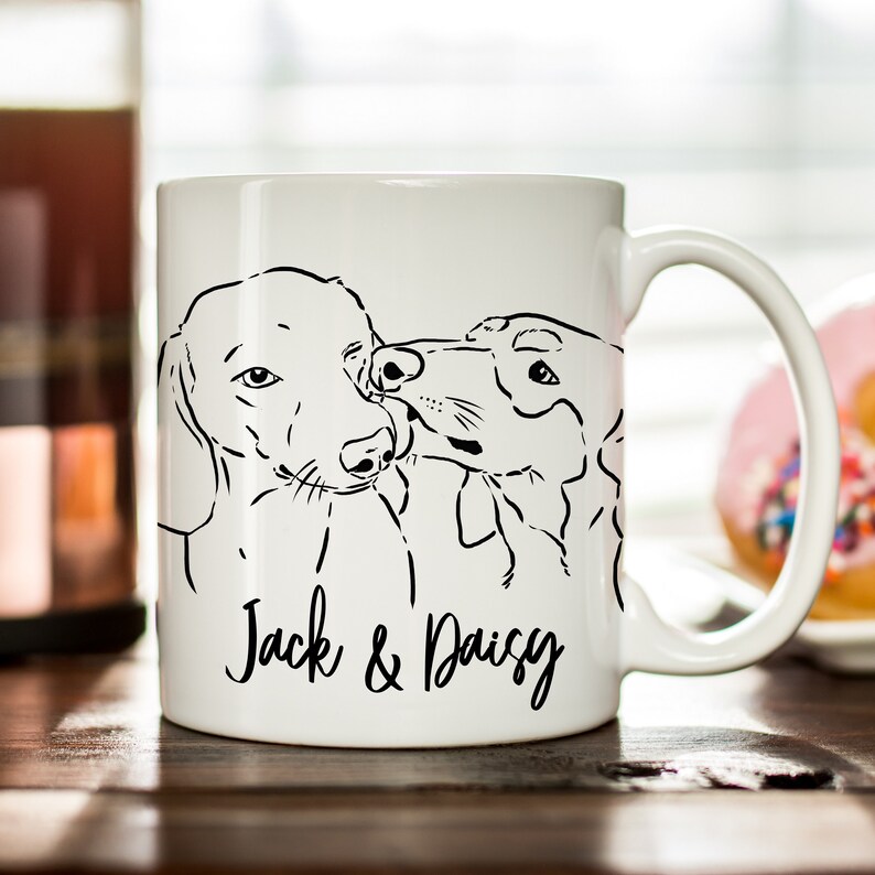 May include: White ceramic mug with a black line drawing of two dogs, one with its head tilted and the other with its head turned to the side, kissing. The text "Jack & Daisy" is written below the dogs.