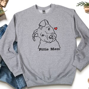 May include: A light gray sweatshirt featuring a black line drawing of a dog's face with a red heart. The text "Pitti Mom" is printed below the dog illustration. The sweatshirt is made of a soft material.
