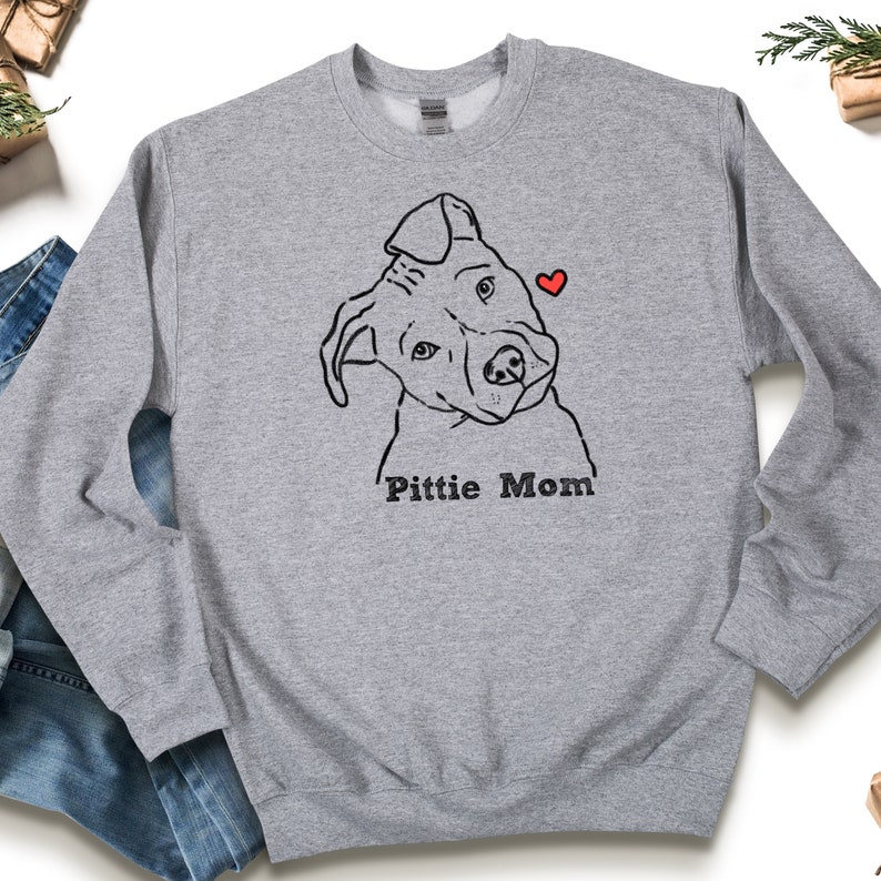 May include: A light gray sweatshirt featuring a black line drawing of a dog's face with a red heart. The text "Pitti Mom" is printed below the dog illustration. The sweatshirt is made of a soft material.