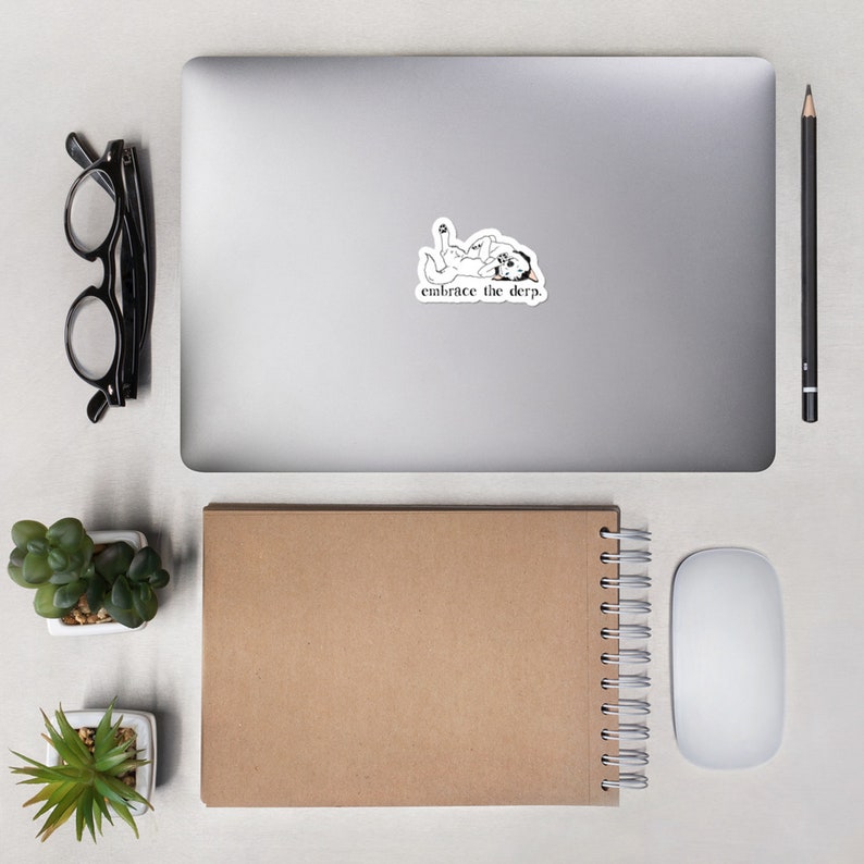 May include: A white laptop computer with a sticker on the lid. The sticker features a black and white illustration of a dog with the text "embrace the derp."