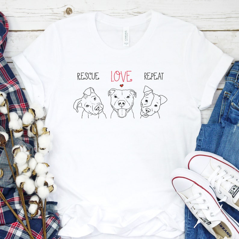 Puede incluir: Camiseta blanca con un dibujo en l&iacute;nea negra de tres perros pitbull. El texto "Rescue Love Repeat" est&aacute; impreso sobre los perros. Un coraz&oacute;n rojo est&aacute; en la palabra "Love".